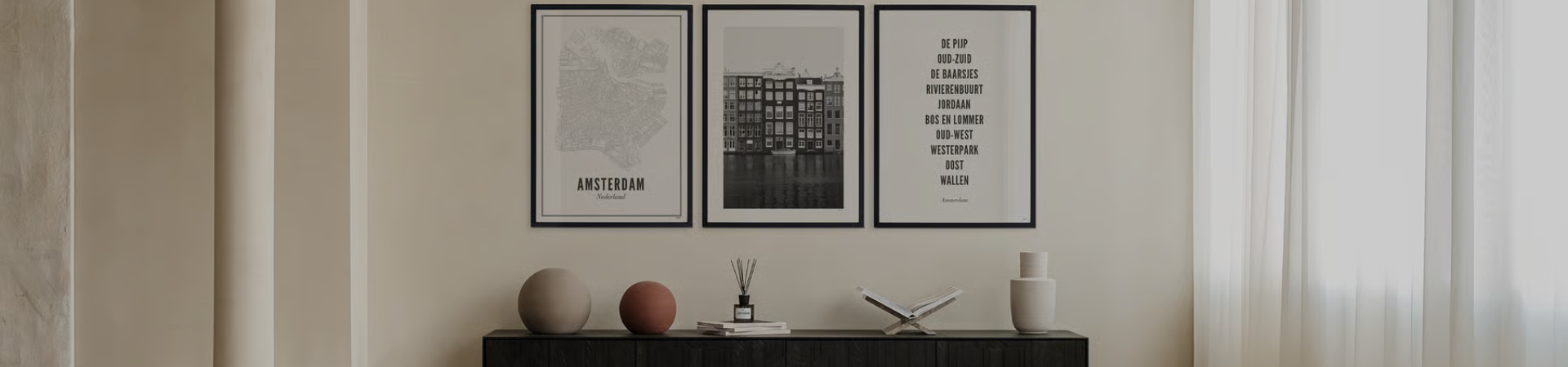 Minimalistische woonkamer met een zwarte consoletafel, twee vazen, een open boek en drie ingelijste kunstdrukken aan de muur erboven, waaronder een kaart, een stadsgezicht en tekst, tegen lichtgekleurde muren en gordijnen.
