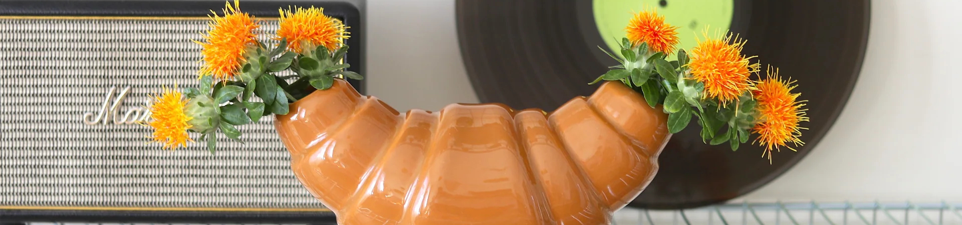 Een keramieken vaas in de vorm van een croissant houdt oranje bloemen vast aan beide uiteinden, met een vintage luidspreker en een vinylplaat op de achtergrond.