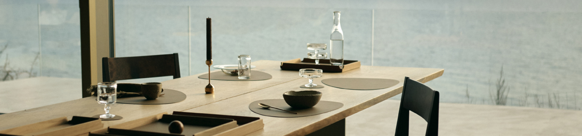 Minimalistische eettafel met zwarte stoelen, bruin en glazen servies, een kaars en een karaf. De grote ramen kijken uit over het serene water buiten.