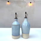 Twee AB Ceramics Olijfolie Fles Wit Opaal 54 cl flessen met metalen tuiten en beige bodems staan naast elkaar op een wit oppervlak, verlicht door twee zachte, warme strijklichtjes op de achtergrond.