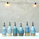 Zes AB Ceramics Olijfolie Fles Grijs Opaal 84 cl met metalen schenktuiten staan op een rij op een wit oppervlak - een stijlvol keukenaccessoire. Boven hangen twee gloeilampen tegen een effen, lichtgekleurde muur.