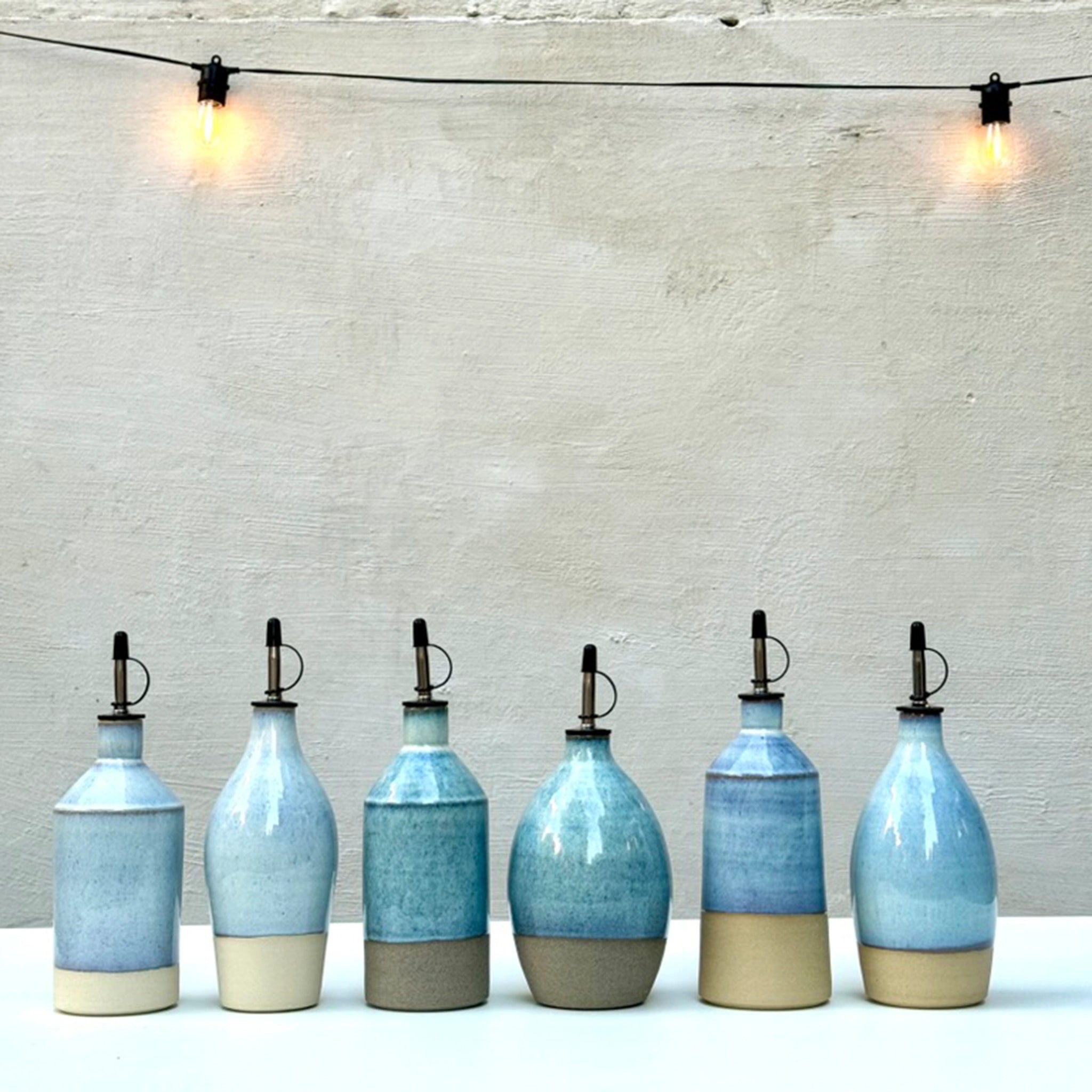 Zes AB Ceramics Olijfolie Fles Grijs Opaal 84 cl met metalen schenktuiten staan op een rij op een wit oppervlak - een stijlvol keukenaccessoire. Boven hangen twee gloeilampen tegen een effen, lichtgekleurde muur.