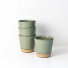 Vier AB Ceramics Beker Geel Groen bekers van 20 cl, gemaakt van steengoed klei met een matgroen glazuur en een ongeglazuurde beige basis, zijn paarsgewijs gestapeld op een wit oppervlak, met één uniek gevormde beker apart vooraan. Merk: AB Ceramics.
