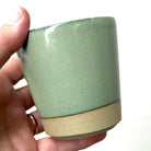 Een hand houdt de AB Ceramics Beker Geel Groen 20 cl van AB Ceramics vast, gemaakt van steengoed klei met een glanzend groengeel glazuur en een ongeglazuurde beige basis, tegen een effen witte achtergrond.