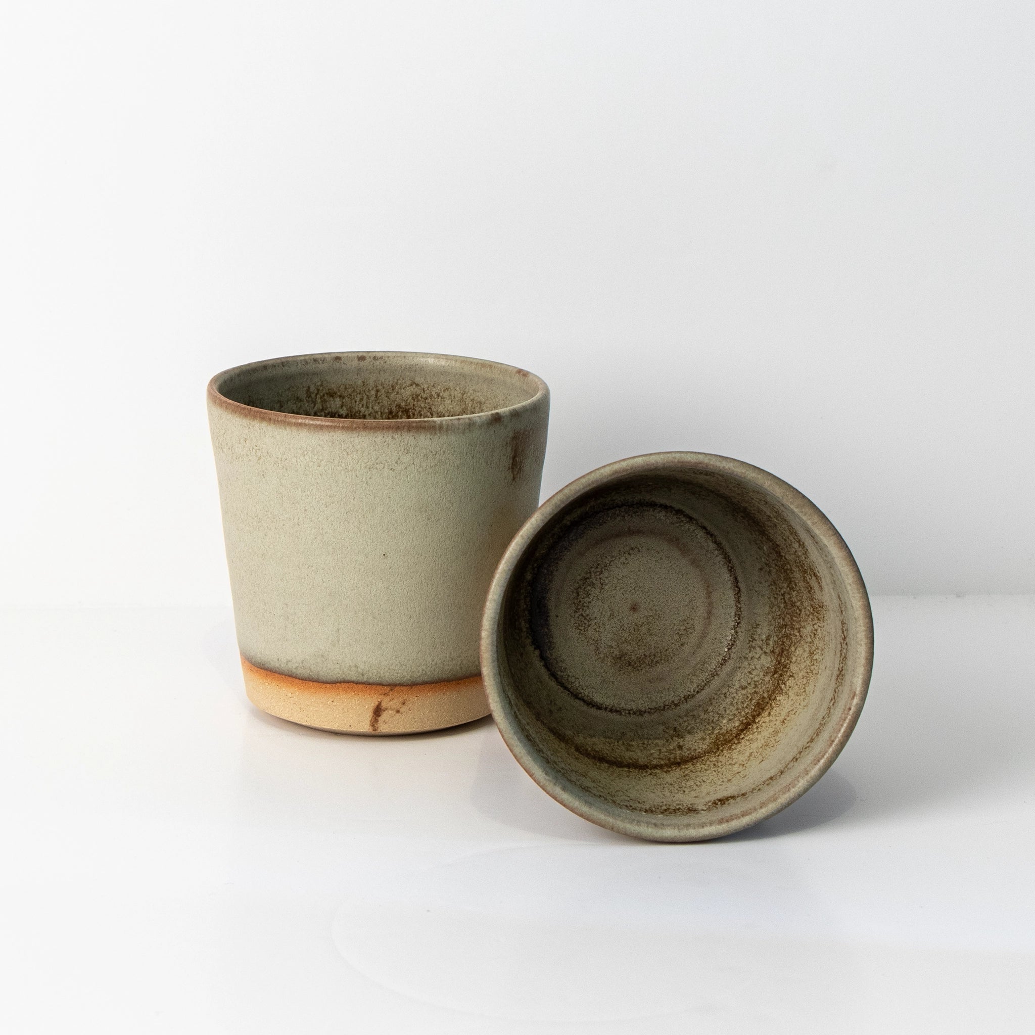 Twee AB Ceramics Beker Geel Mos 20cl van steengoed met een matte, aardse glazuur. Eén beker staat rechtop, de ander ligt op zijn zij en toont het cirkelpatroon binnenin. De effen witte achtergrond benadrukt het ontwerp.