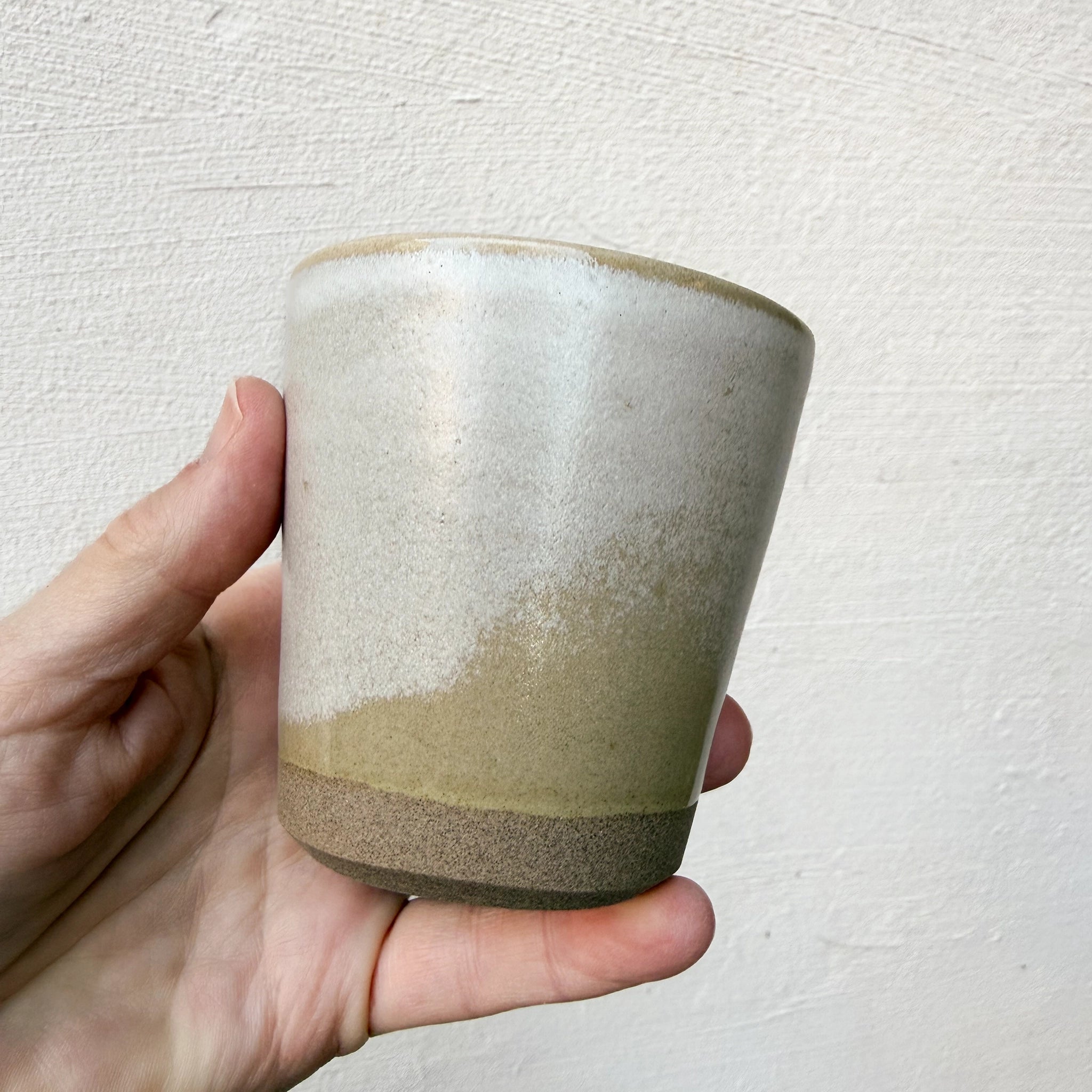 Een hand houdt de AB Ceramics Beker Toffee Satijn 20 cl vast - een kleine mok met een matte afwerking en een kleurverloop van lichtbeige naar aardebruin - tegen een gestructureerde witte achtergrond.