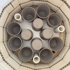 Bovenaanzicht van een oven met handgegoten, ongeglazuurde kopjes van klei in een cirkel. Drie AB Ceramics Beker Toffee Satijn bekers van 20 cl staan er gelijkmatig tussen. De binnenwanden van de oven zijn bekleed met lichtgekleurde bakstenen.