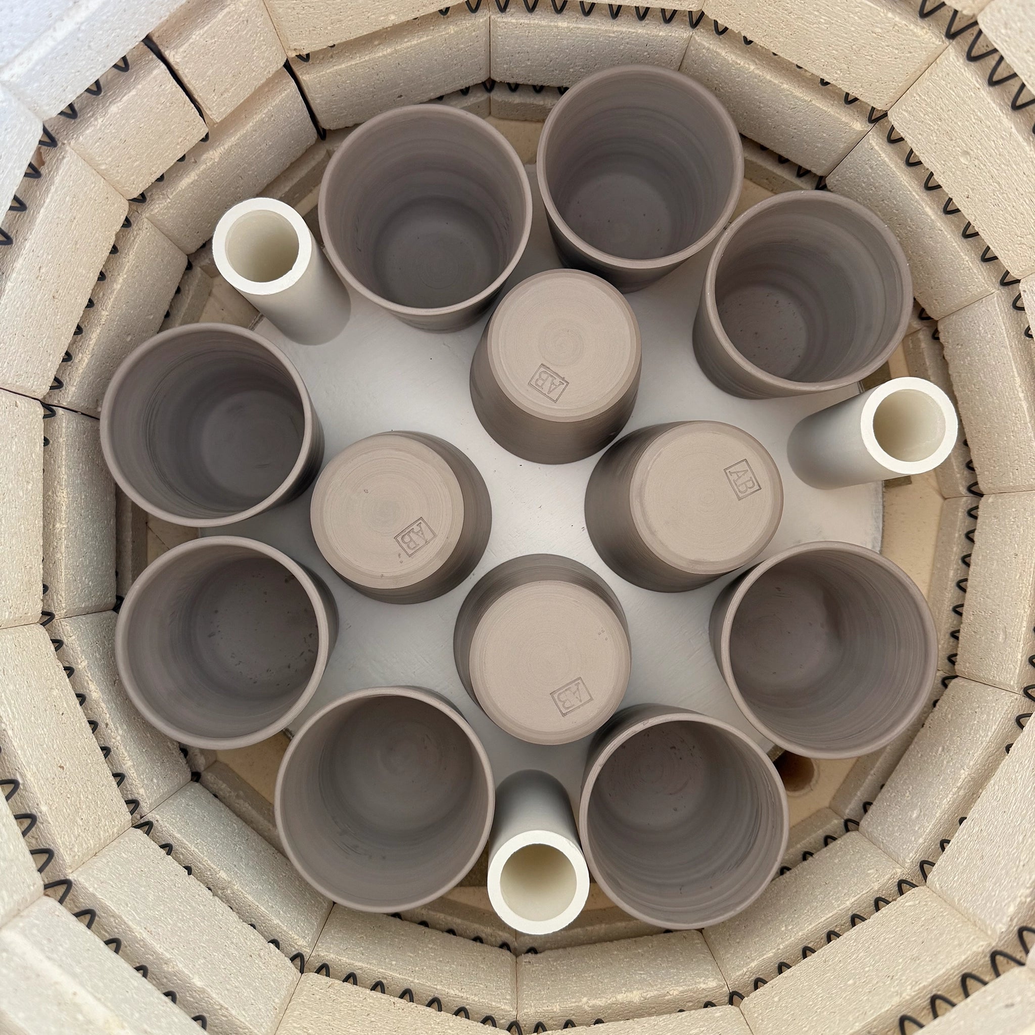 Bovenaanzicht van een oven met handgegoten, ongeglazuurde kopjes van klei in een cirkel. Drie AB Ceramics Beker Toffee Satijn bekers van 20 cl staan er gelijkmatig tussen. De binnenwanden van de oven zijn bekleed met lichtgekleurde bakstenen.