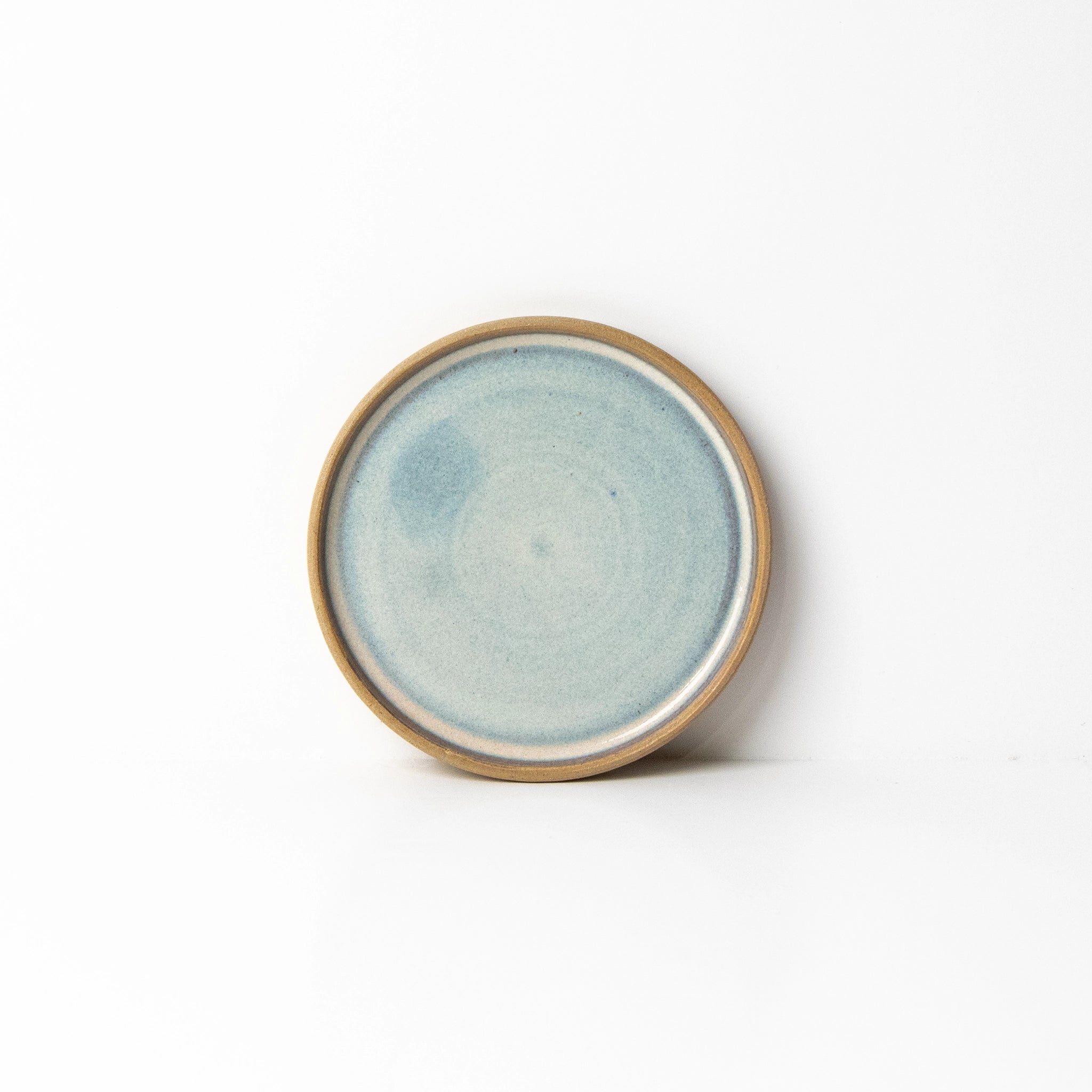 Het AB Ceramics Bordje Mini Geel Opaal/Blauw 12 cm is een handgegoten rond bord van keramiek met een lichtblauw geglazuurd midden en bruine rand, gemaakt van steengoedklei en afgebeeld op een effen witte ondergrond.