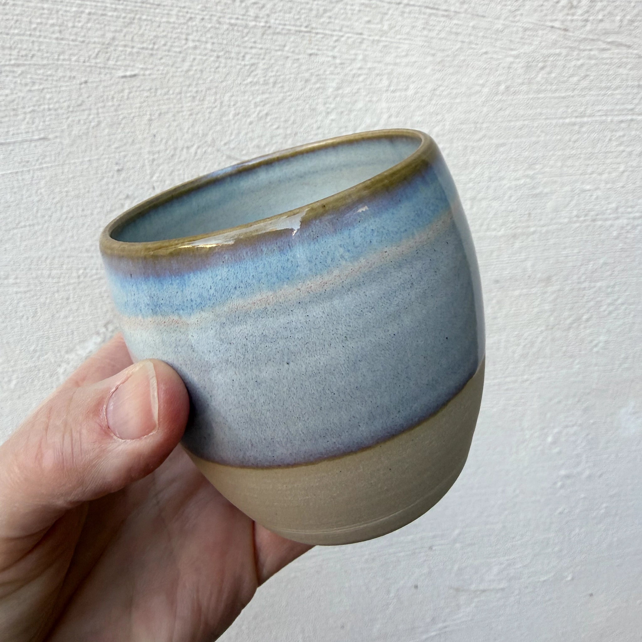 Een hand houdt de AB Ceramics Mok Wit Opaal/Blauw vast - een ronde steengoed mok van AB Ceramics, met een glad, mat glazuur in beige, blauw en bruin tegen een gestructureerde witte achtergrond.