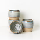Vier AB Ceramics Mok Wit Opaal/Blauw steengoed mokken, handgedraaid en gestapeld met een lichtblauw-grijs naar beige glazuur. Eén mok ligt op zij en toont het unieke keramiek interieur van AB Ceramics.