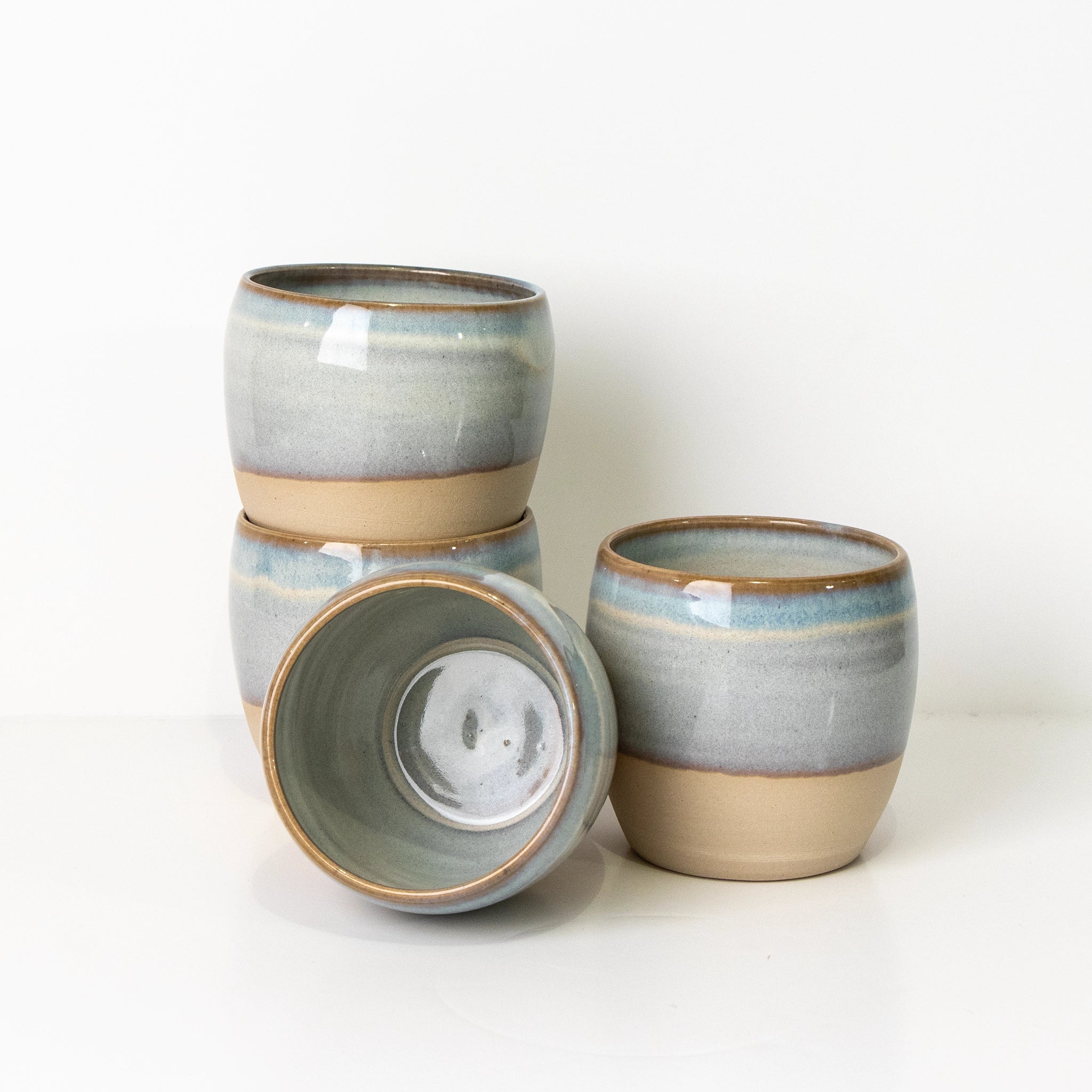 Vier AB Ceramics Mok Wit Opaal/Blauw steengoed mokken, handgedraaid en gestapeld met een lichtblauw-grijs naar beige glazuur. Eén mok ligt op zij en toont het unieke keramiek interieur van AB Ceramics.