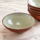 De AB Ceramics Noodle Kom Rood Mint D19 heeft een matbruine buitenkant en glanzende lichtgroene binnenkant, handgemaakt door AB Ceramics. Afgebeeld rustend op marmer naast vergelijkbare kommen.