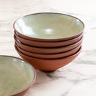 Een stapel van vijf AB Ceramics Noodle Kom Rood Mint D19 kommen staat op een wit marmeren oppervlak, met één bijpassende kom apart ervoor.