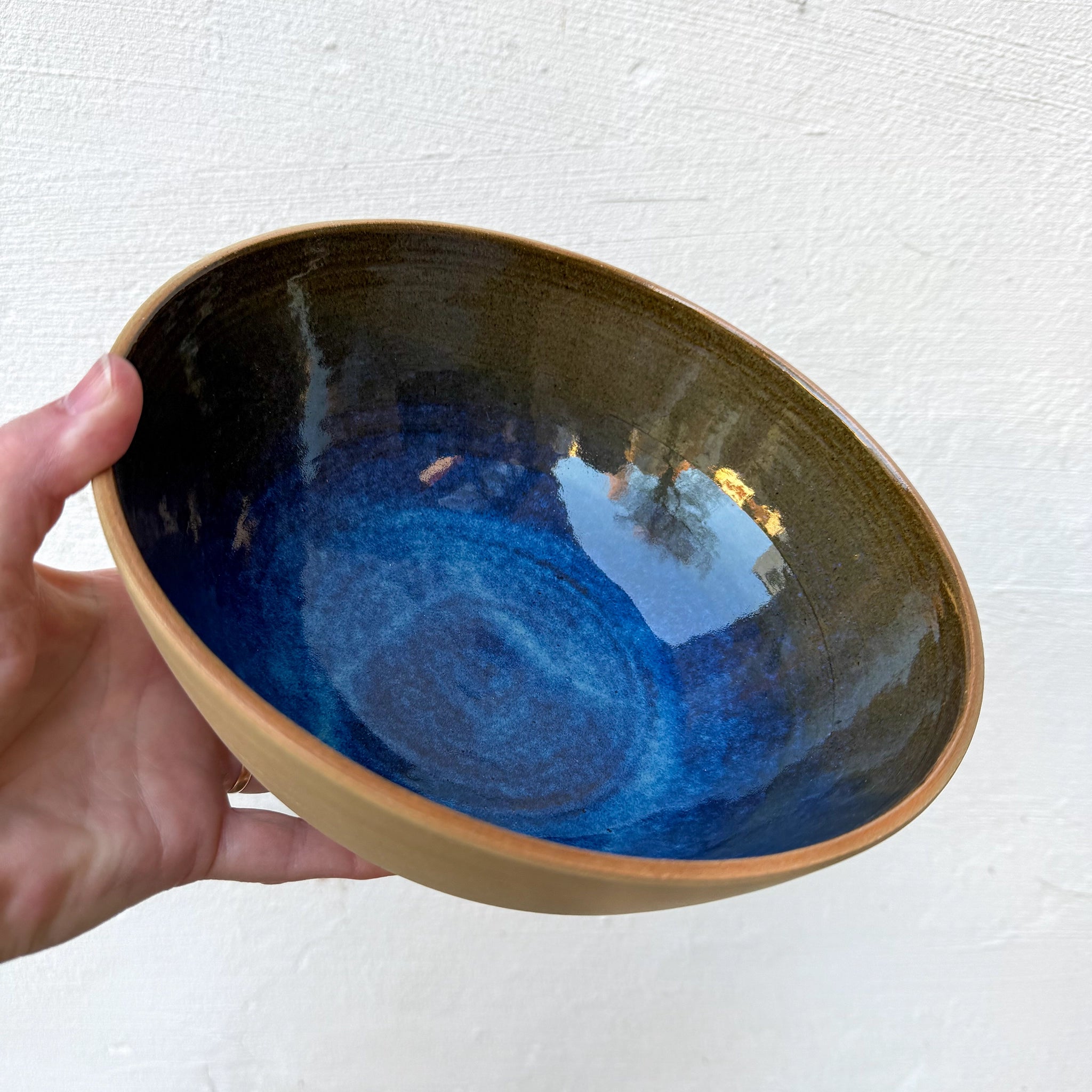 Een hand houdt de AB Ceramics Serveerschaal Geel Blauw D23 van AB Ceramics vast, met een matgele buitenkant en glanzend diepblauwe binnenkant, die licht weerkaatst tegen een effen gebroken witte muur.