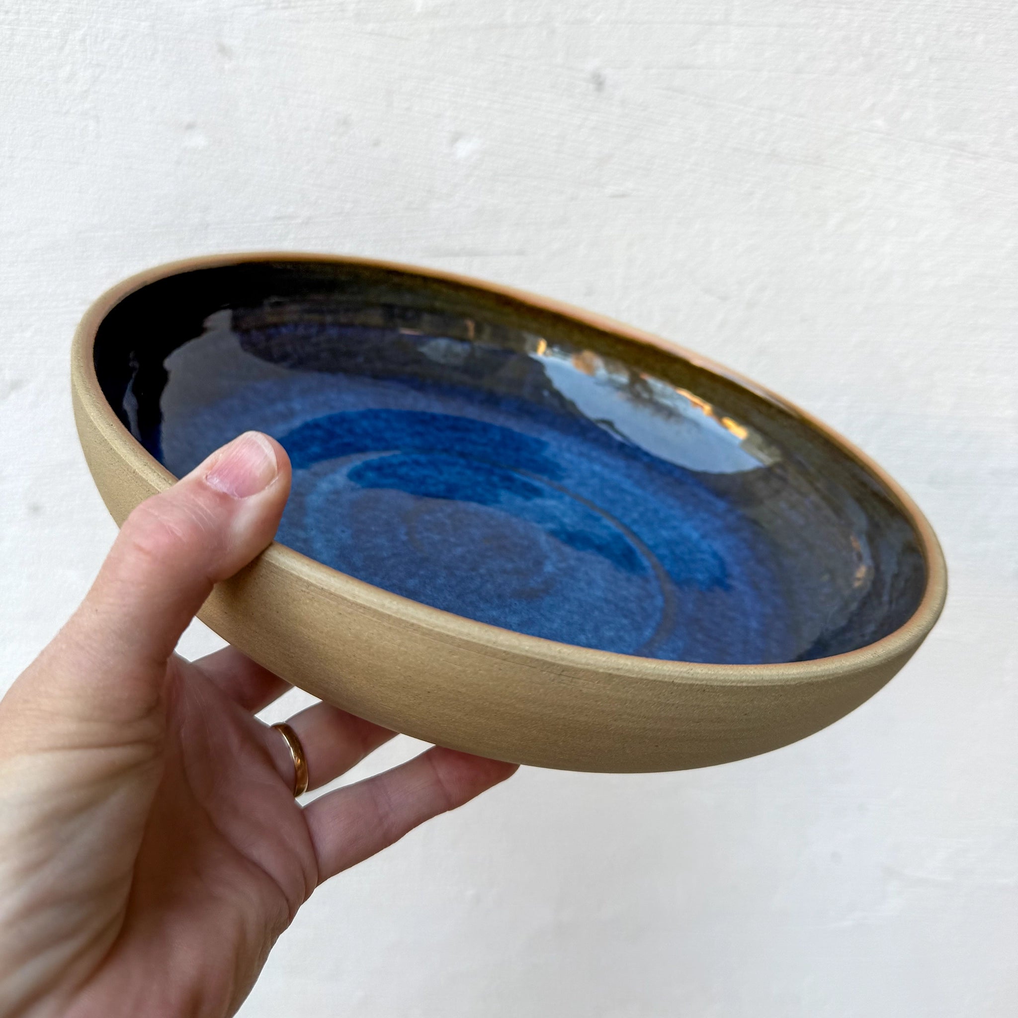 Een hand houdt de AB Ceramics Serveerschaal Geel Blauw D25 vast - de matte tan steengoed buitenkant contrasteert met een glanzende blauwe binnenkant - tegen een effen, lichte achtergrond.