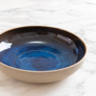 De AB Ceramics Serveerschaal Geel Blauw D25, een handgedraaide schaal van gele steengoed klei met beige buitenkant en glanzend diepblauw interieur, staat op een wit marmeren oppervlak.