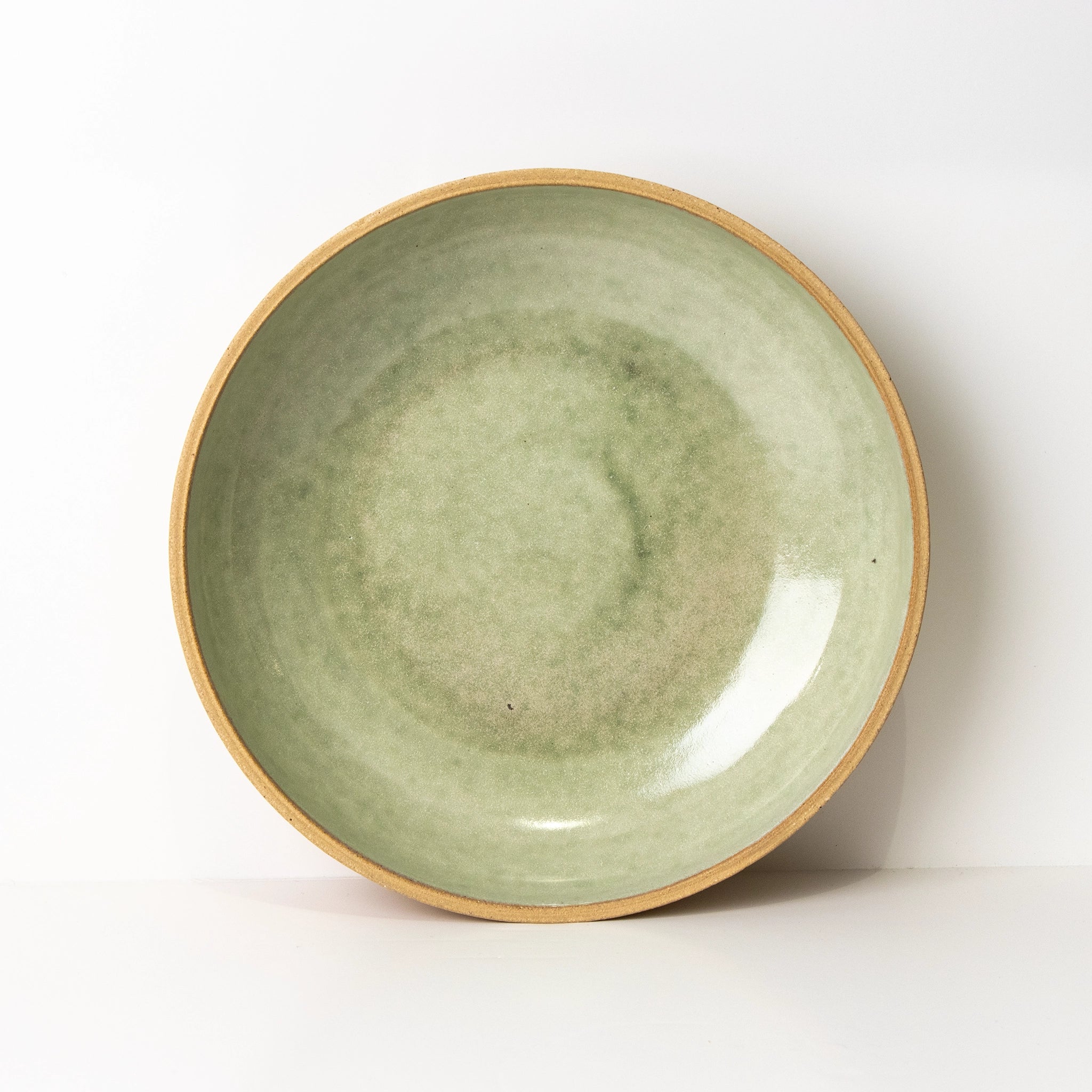 De AB Ceramics Serveerschaal Ruw Mint D27,5, een ronde steengoed schaal met mintkleurig glazuur en een ongeglazuurde rand, staat rechtop tegen een witte achtergrond.