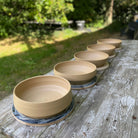 Zes AB Ceramics Serveerschalen Ruw Mint D25 staan op een verweerde houten buitentafel, omringd door groene bladeren en zacht zonlicht.