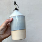 Een hand houdt de AB Ceramics Olijfolie Fles 1 Prai Opaal 65 cl vast tegen een grijze muur, zodat je de puntige schouder en het AB-stempel goed ziet.
