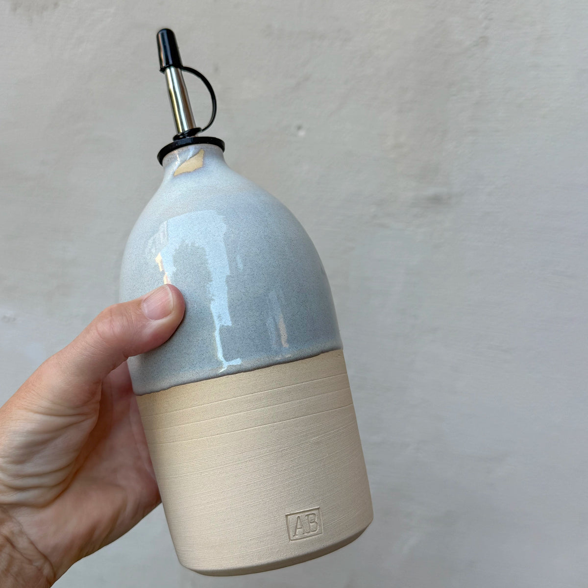Een hand houdt de slanke AB Ceramics Olijfolie Fles 3 Prai Opaal 65 cl vast tegen een grijze muur.