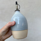 Een hand houdt de eivormige AB Ceramics Olijfolie Fles 4 Prai Opaal 65 cl vast tegen een grijze muur.