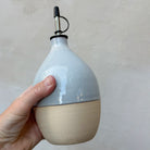 Een hand houdt de peervormige AB Ceramics Olijfolie Fles 5 Prai Opaal 70 cl vast tegen een grijze muur.
