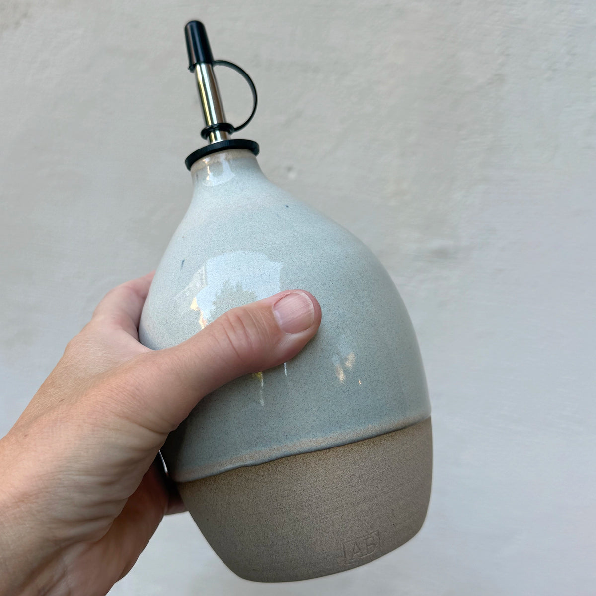 Een hand houdt de AB Ceramics Olijfolie Fles 6 Toffee Opaal 70 cl vast tegen een grijze muur.