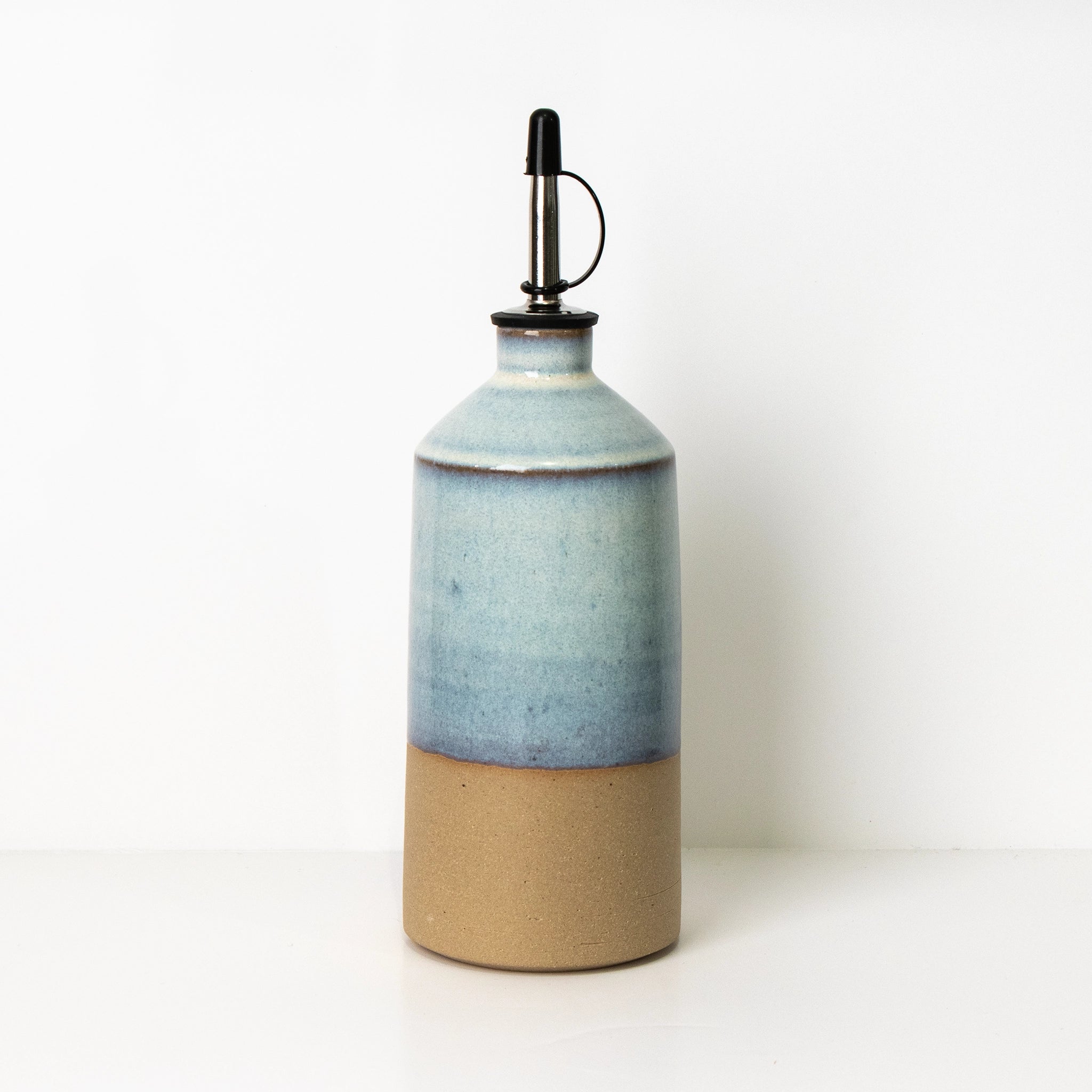 De AB Ceramics Olijfolie Fles Geel Opaal 60 cl van AB Ceramics is een oliedispenser van keramiek met een beige onderkant, een blauw verloop boven en een metalen tuitdeksel, afgebeeld op een effen witte achtergrond.