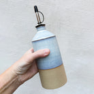 Een hand houdt een AB Ceramics Olijfolie Fles Geel Opaal 60 cl vast, met een geel opaalglazuur aan de bovenkant en natuurlijk beige aan de onderkant, met een metalen tuit, tegen een licht gestructureerde achtergrond.