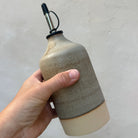 Een hand houdt de AB Ceramics Olijfolie Fles Recht Wit Mos 55 cl van AB Ceramics vast, met aardebeige en grijze keramiektinten, een metalen tuit met zwarte rubberen stop, tegen een effen lichte achtergrond.