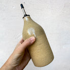 Een hand houdt een AB Ceramics Olijfolie Fles Ruw Transparant 60 cl vast, een beige keramische fles met metalen tuit van AB Ceramics, ideaal voor het schenken van olie of azijn, tegen een lichte achtergrond.