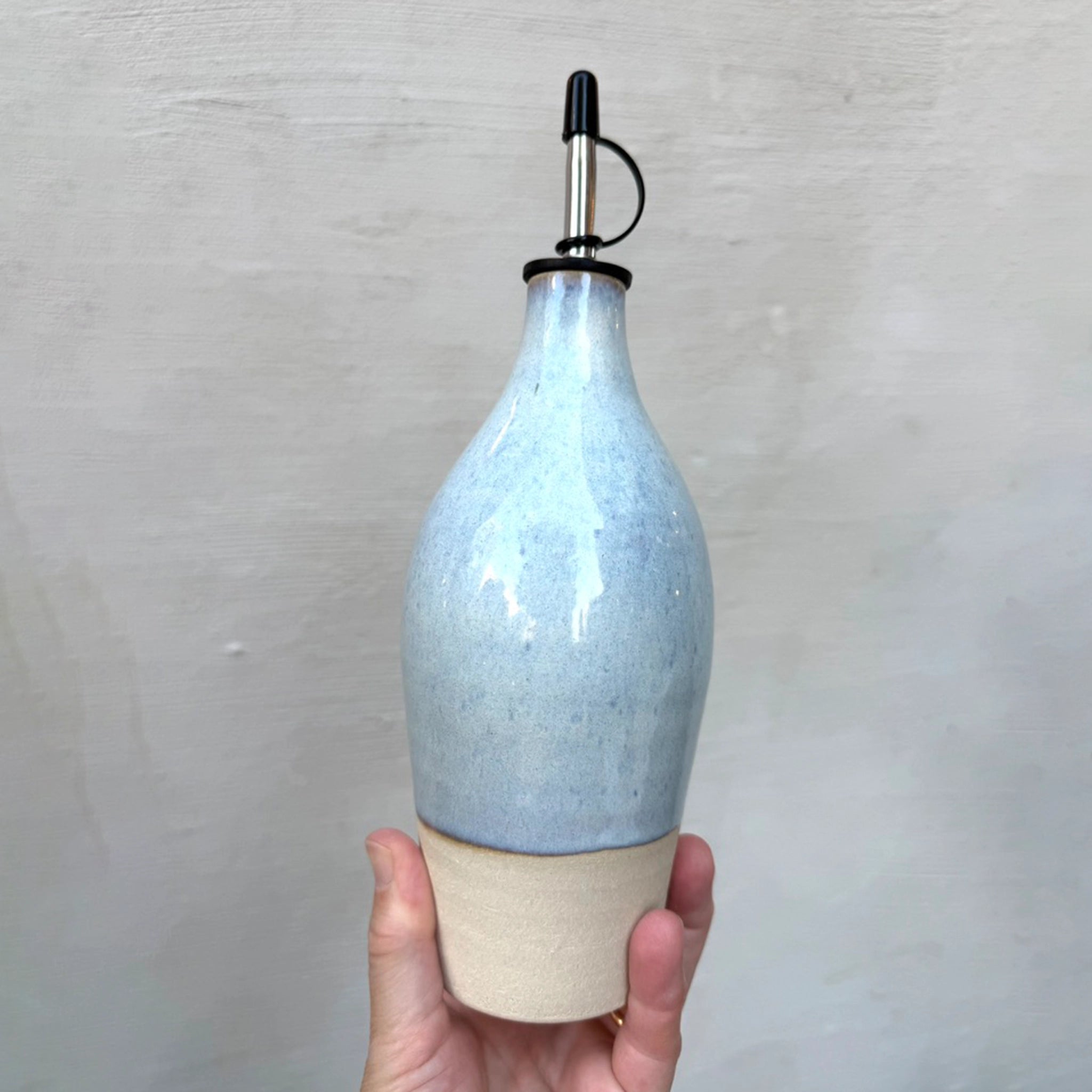 Een hand houdt de AB Ceramics Olijfolie Fles Wit Opaal 54 cl vast, een handgemaakte oliefles van keramiek met een lichtblauw opaal geglazuurde bovenkant, natuurlijke beige basis en metalen tuit met zwarte rubberen dop tegen een neutrale achtergrond.