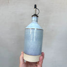 Een hand houdt de AB Ceramics Olijfolie Fles Wit Opaal 56 cl met glanzend lichtblauw glazuur, beige basis en metalen tuit met zwarte dop vast - een elegant stuk van AB Ceramics dat perfect is voor je keukenaccessoires.