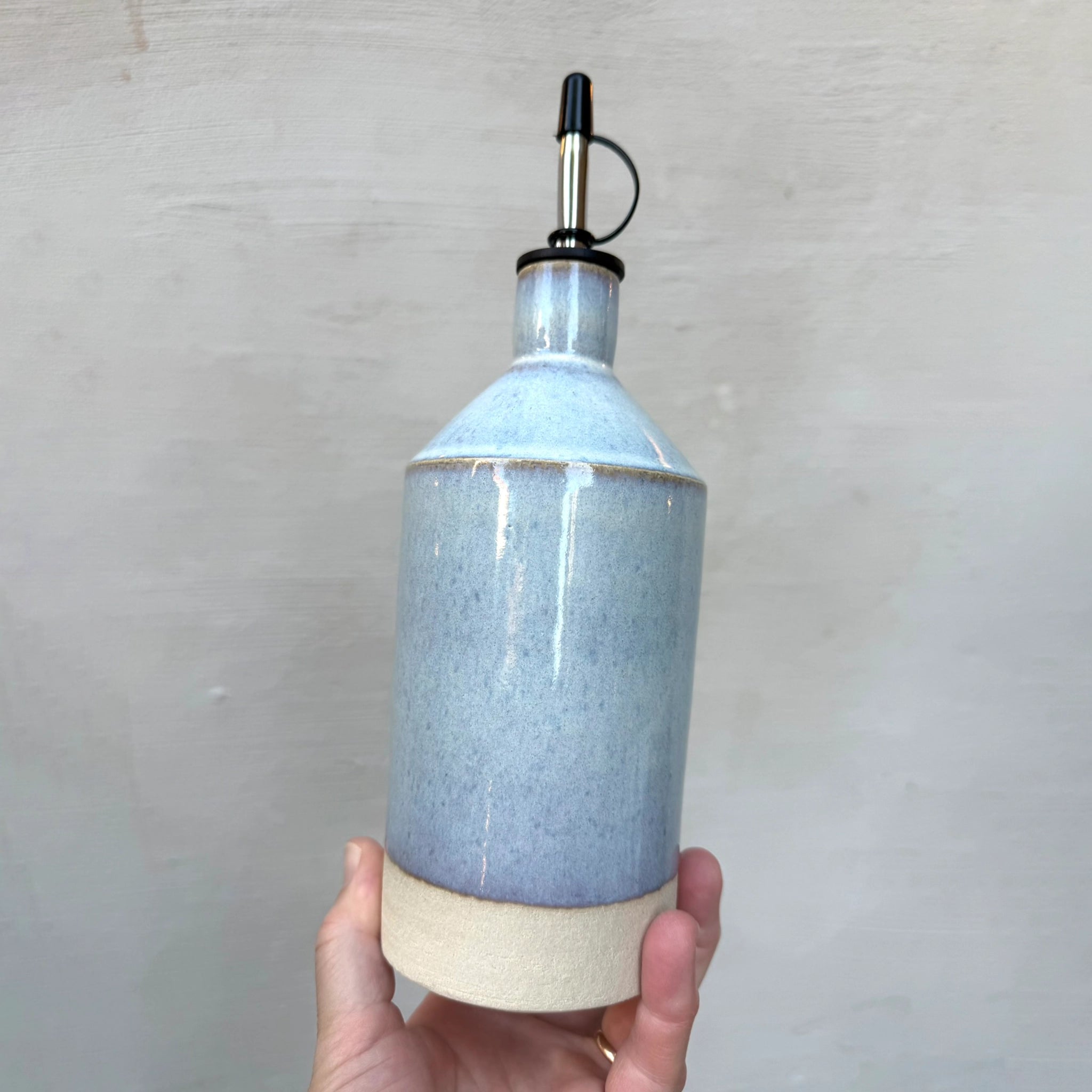 Een hand houdt de AB Ceramics Olijfolie Fles Wit Opaal 56 cl met glanzend lichtblauw glazuur, beige basis en metalen tuit met zwarte dop vast - een elegant stuk van AB Ceramics dat perfect is voor je keukenaccessoires.