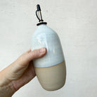 Een hand houdt de AB Ceramics Olijfolie Fles Wit Opaal 70 cl vast, met een matbeige basis, glanzend witte bovenkant en een metaal-rubberen tuit, tegen een licht getextureerde achtergrond.