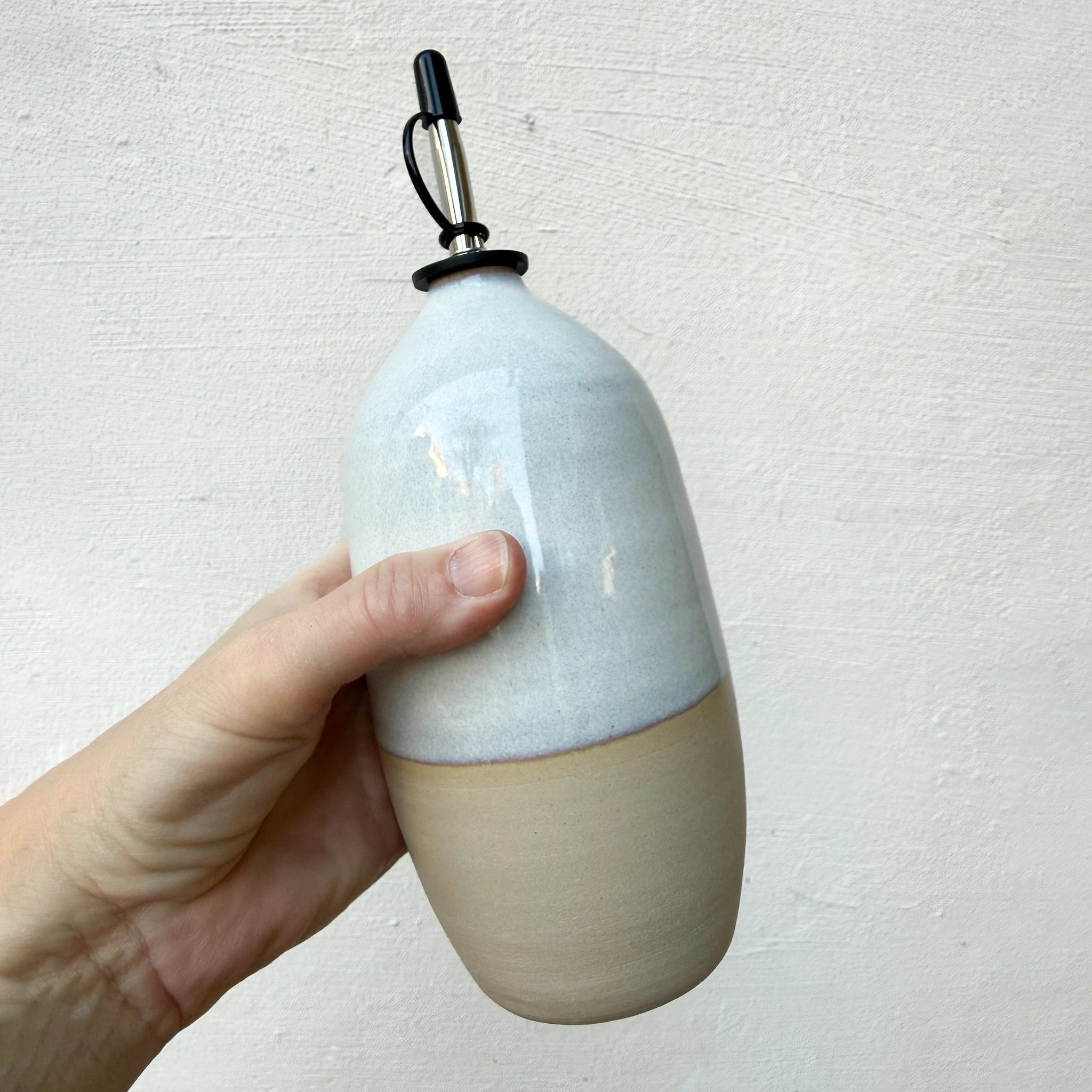 Een hand houdt de AB Ceramics Olijfolie Fles Wit Opaal 70 cl vast, met een matbeige basis, glanzend witte bovenkant en een metaal-rubberen tuit, tegen een licht getextureerde achtergrond.