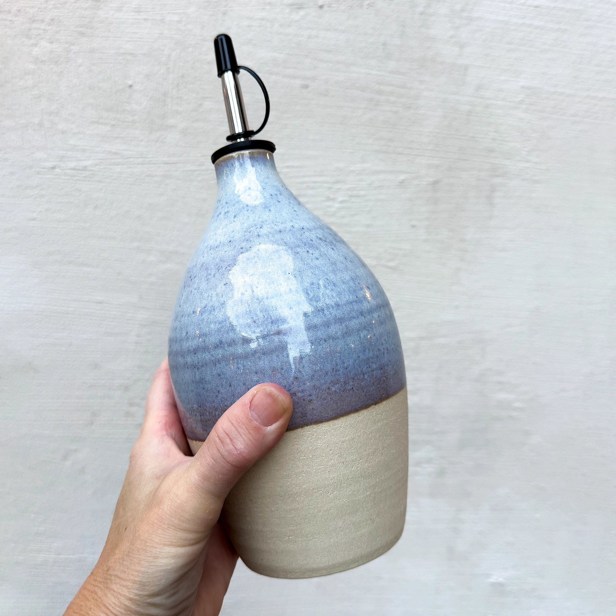Een hand houdt de AB Ceramics Olijfolie Fles Wit Opaal 85 cl vast, met een matbeige basis en glanzende lichtblauwe bovenkant met een metalen schenktuit en zwarte dop - een opvallende AB Ceramics keukenaccessoire tegen een effen achtergrond.