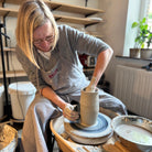 Een vrouw met een bril en een schort vormt de AB Ceramics Olijfolie Fles Grijs Opaal 84 cl op een pottenbakkersschijf - een uniek keukenaccessoire - omringd door planken en kunstbenodigdheden in een lichte studio.