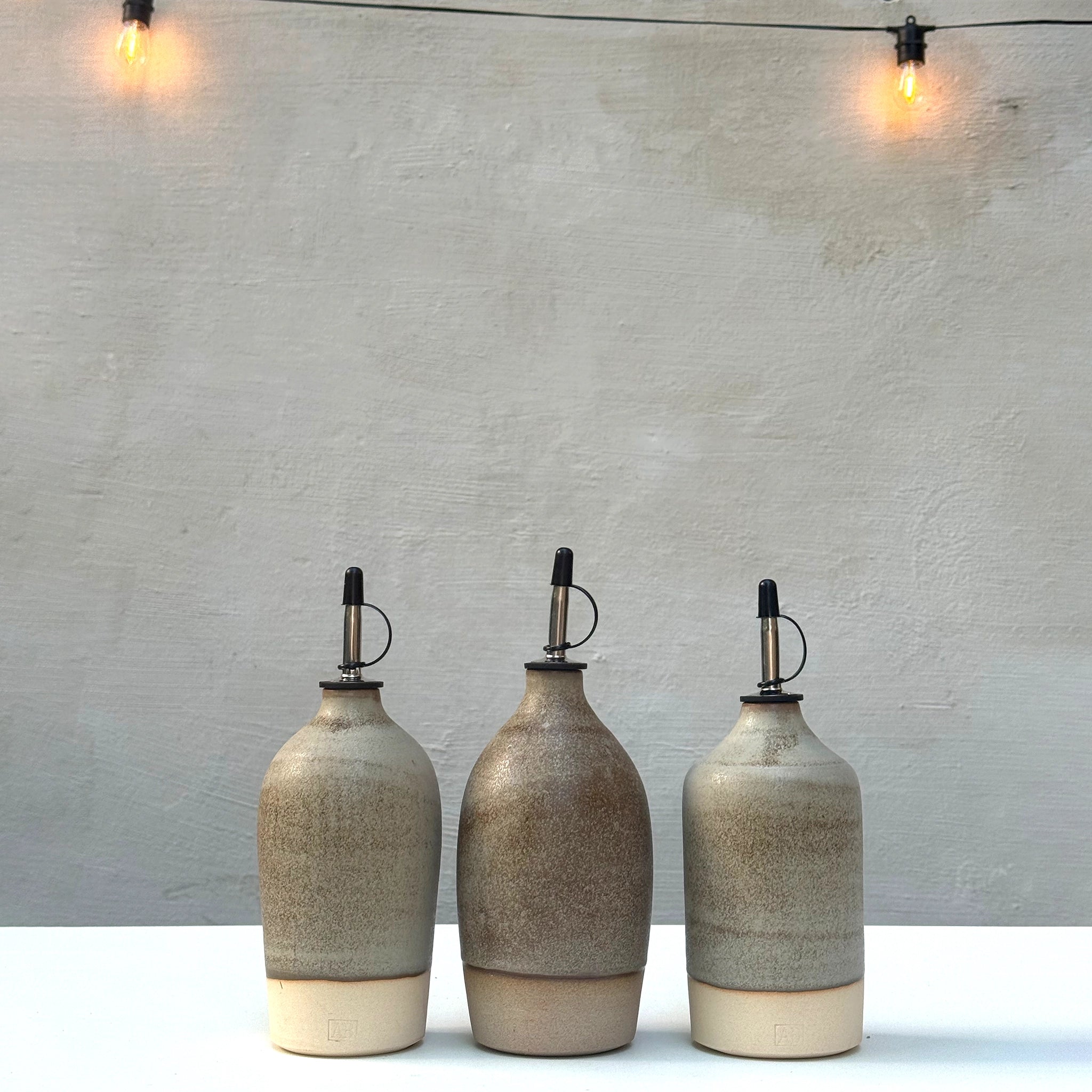 Drie AB Ceramics Olijfolie Fles 2 Wit Mos 55 cl staan op een rij op een witte ondergrond voor een grijze muur, met warme string lights erboven-een stijlvolle accessoire voor elke keuken.