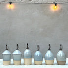 Zes handgedraaide AB Ceramics olijfolieflessen, elk met een andere vorm, op een witte tafel tegen een grijze muur met twee brandende lampjes.
