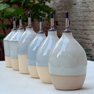 Een rij handgedraaide AB Ceramics olijfolieflessen met schenktuit buiten op een witte tafel, met groen op de achtergrond.