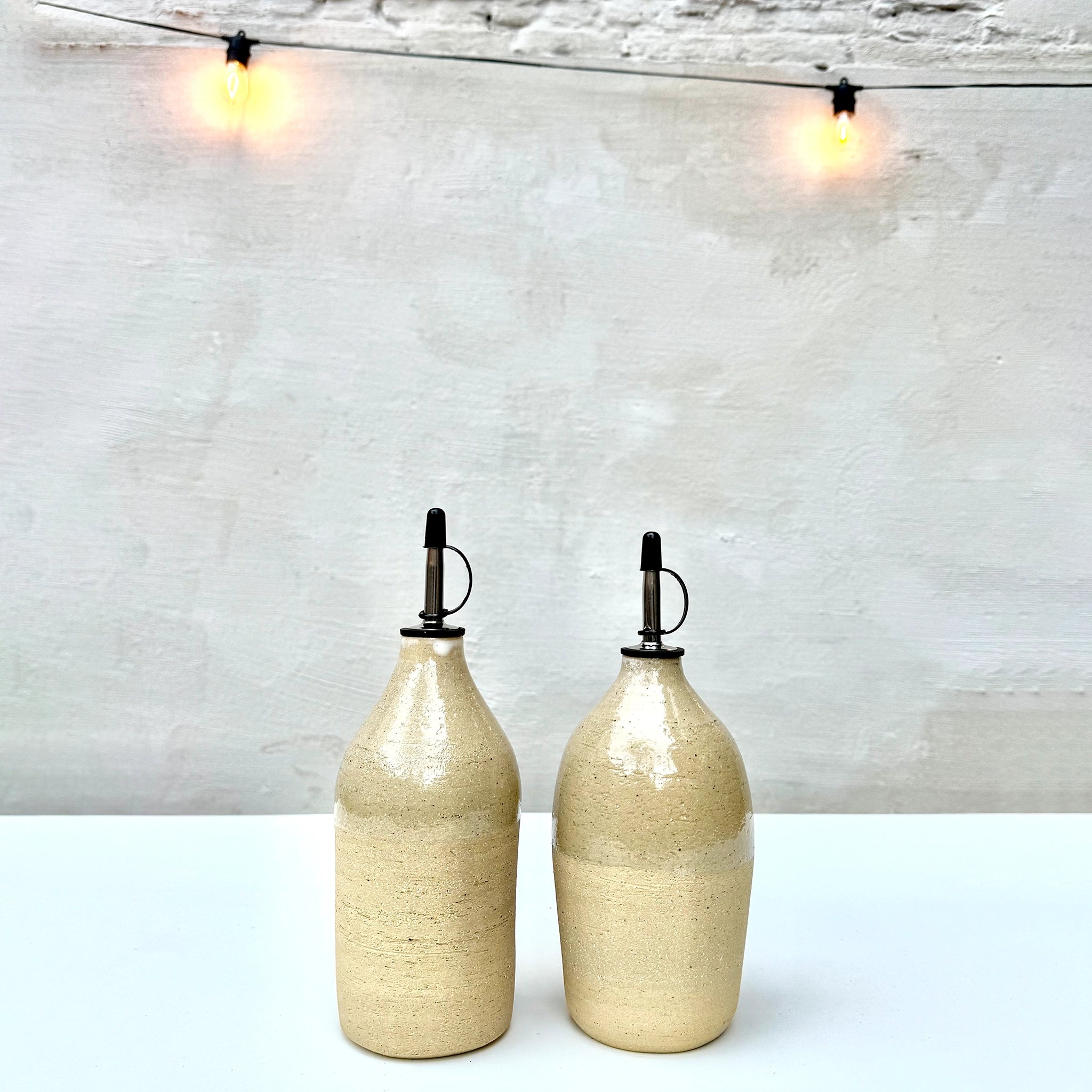 Twee AB Ceramics Olijfolie Fles Ruw Transparant 60 cl dispensers met metalen tuiten staan op een wit oppervlak voor een lichtgrijze muur, verlicht door warme strijklichtjes die de kenmerkende stijl van AB Ceramics oproepen.