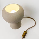 De AB Ceramics Sfeerlamp 20cm van AB Ceramics is een kleine, ronde lamp van beige keramiek met een getextureerde afwerking, gouden aan/uit-schakelaar aan een gevlochten bruin snoer en straalt zacht warm licht uit vanuit de open bovenkant.