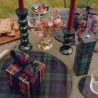 Op een buitentafel liggen geruite placemats, ingepakte cadeautjes, een 24 cm Addison Ross Bobbin Kandelaar in Racing Green met rode kaarsen, een vaas met wilde bloemen, glaswerk en een houten plank met sneetjes brood.