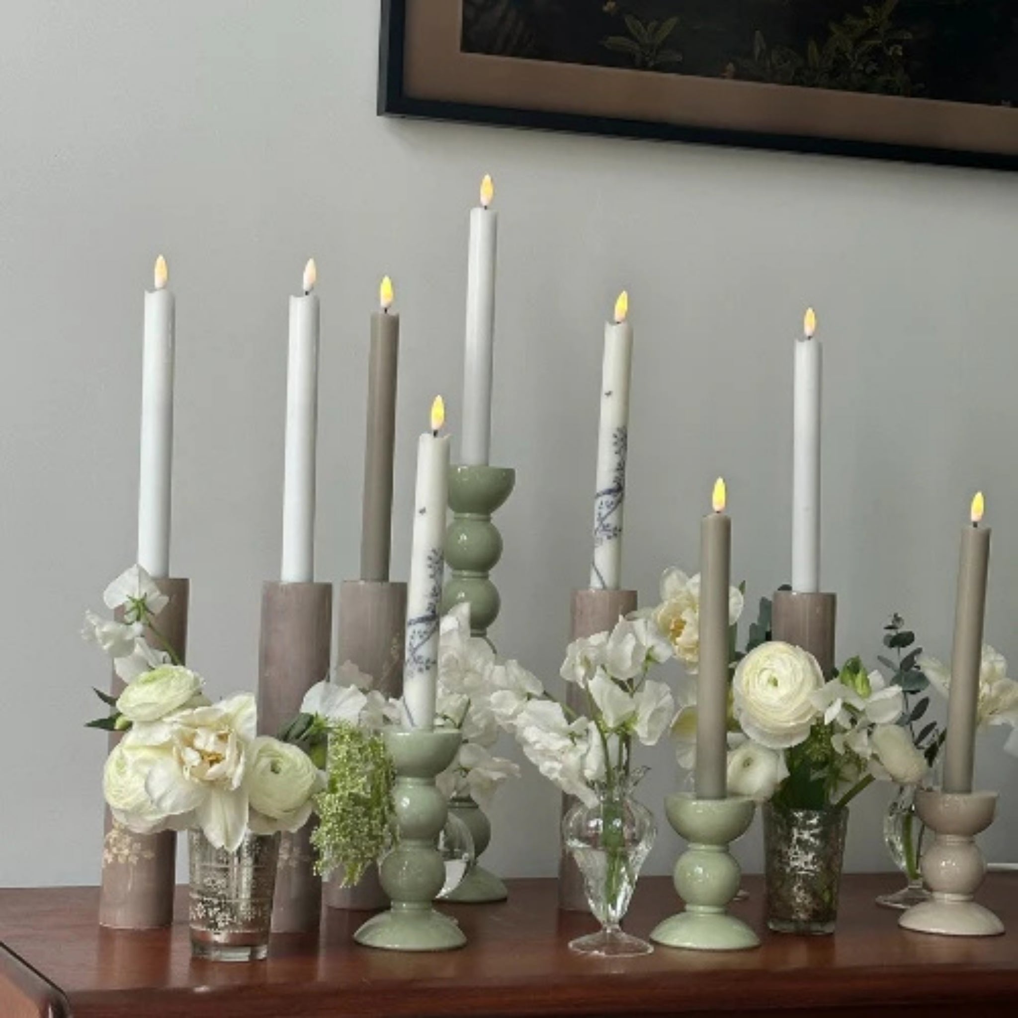 Een groep witte en grijze taps toelopende kaarsen in keramische houders, waaronder de Addison Ross Bobbin Kandelaar Saliegroen 10 cm, met verschillende vazen met witte bloemen zijn gerangschikt op een houten oppervlak tegen een lichte muur. Sommige kaarsen gloeien warm op.