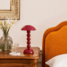 Op een houten nachtkastje staan een vaas met witte bloemen, een figuurglas met een schoteltje en de Addison Ross Bobbin Lamp LED Oplaadbaar Kersenrood; een deel van een oranje gestoffeerd bed en een goudomrande spiegel zijn ook zichtbaar.