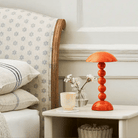 Op een nachtkastje naast het bed staan een Addison Ross Bobbin Lamp LED Oplaadbaar Oranje, een glas water, witte bloemen in een kleine vaas en een kaars. Gedessineerde kussens sieren het bed en het tafeltje staat tegen een witte muur.