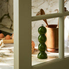 Een Addison Ross Bobbin Zout-en Pepermolen Kaki 24 cm staat op een marmeren aanrecht in de buurt van een houten keukengereihouder, snijplank met eieren, knoflook en een ui, allemaal gezien door een raamkozijn.