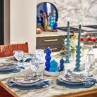 Op een marmeren keukentafel voor vier personen staan blauwe en witte borden, kobaltkleurig glaswerk, blauwe kandelaars met hoge kaarsen, een middenstuk van gestapelde blauwe glazen bollen, een Addison Ross Bobbin Zout-en Pepermolen Kobalt 24 cm en verse bloemen.